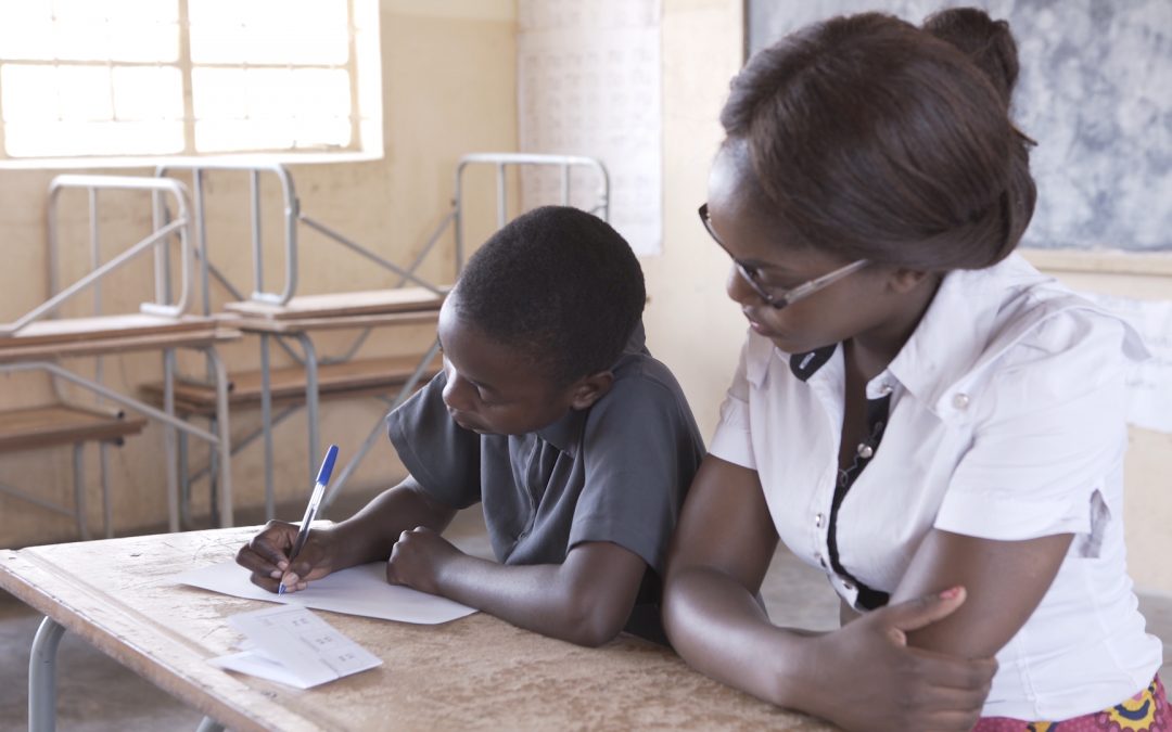 A child completes a sum on a piece of paper while a teacher watches during a TaRL assessment in Zambia.