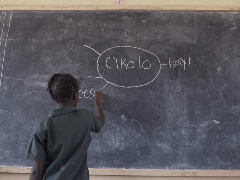 A child writes the word cesu on a mind map on a board. In the centre of the mind map is the word "cikolo".