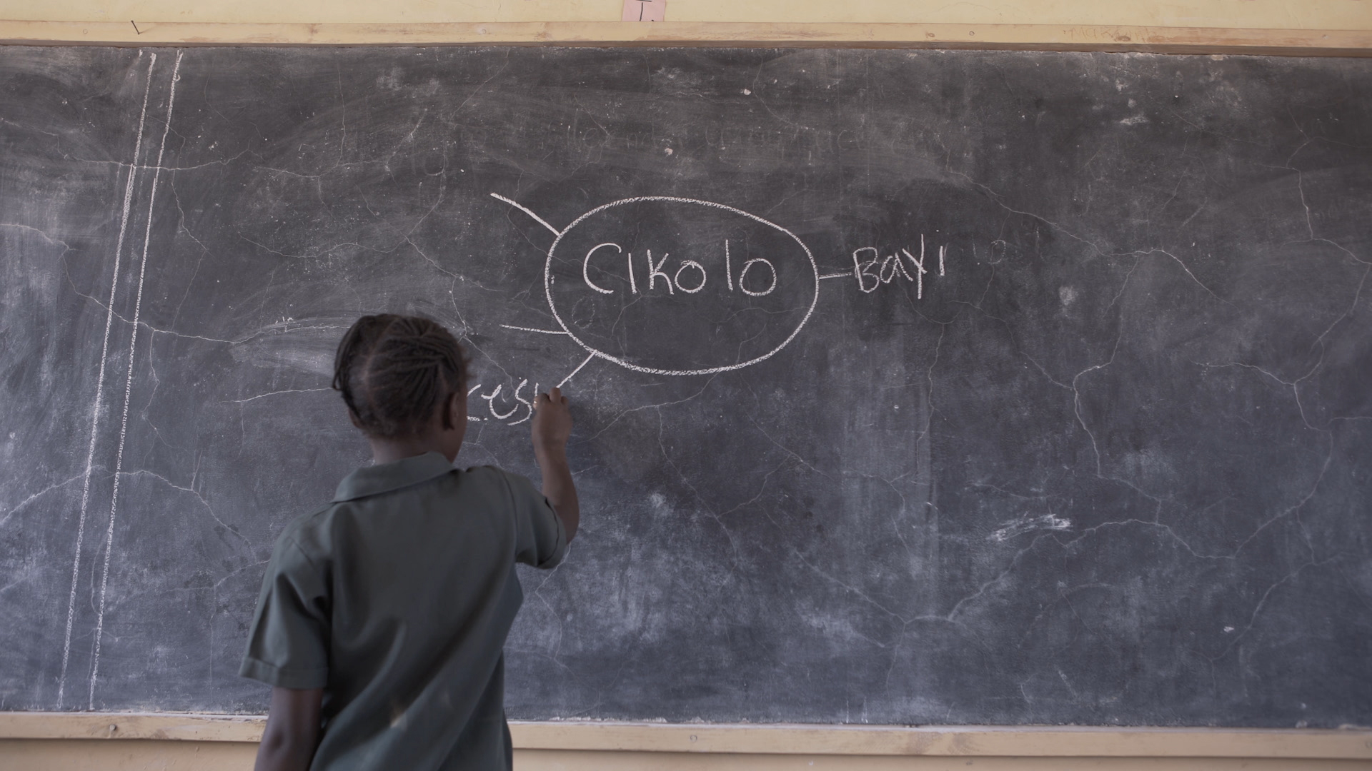 A child stands at the board, writing the word "cesu" in chalk on a mind map during a TaRL reading class in Zambia. On the board, the word "cikolo" is circled, with lines to "bayi" and "cesu".