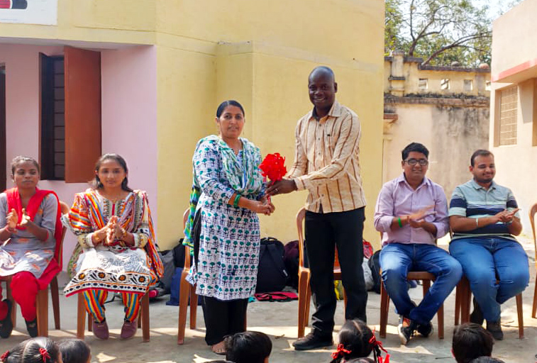 A woman gives a big red flower to a man. People sit behind them on chairs, school children sitting in front of them on the floor.