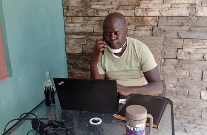 Richard Bwale sits at a table holding a cellphone to his ear, looking at a laptop on the desk in front of him, a medical mask around his neck.