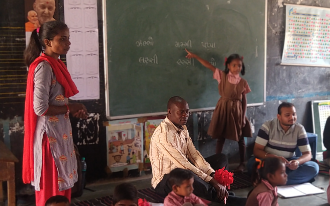 A teacher stands in a classroom, a child stands and points at the board. A group of children and two adults sit on the floor in front of the board.